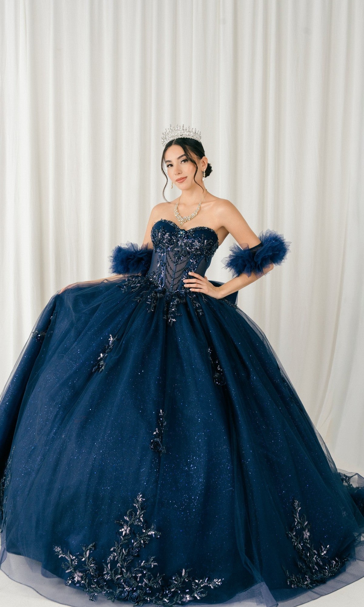 A young woman wears the Navy Quinceanera Dress 8380J By Cinderella Couture with matching arm cuffs, a tiara, and jewelry as she stands before a white curtain backdrop.